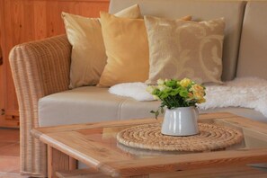 A wicker sofa with beige cushions and a white throw blanket, a glass coffee table with a woven placemat, and a vase with yellow and green flowers. - "Am Teich" on the Sanderhof (Rathmannsdorf)