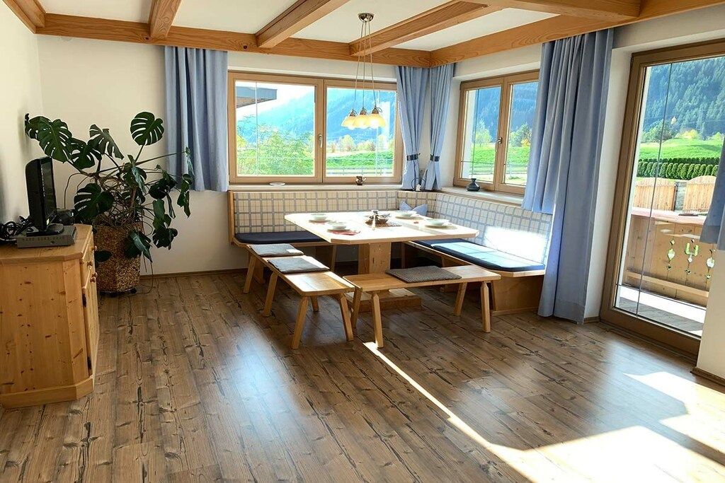 Une pièce avec un plancher en bois, une table à manger avec des bancs, une plante en pot et de grandes fenêtres avec des rideaux.