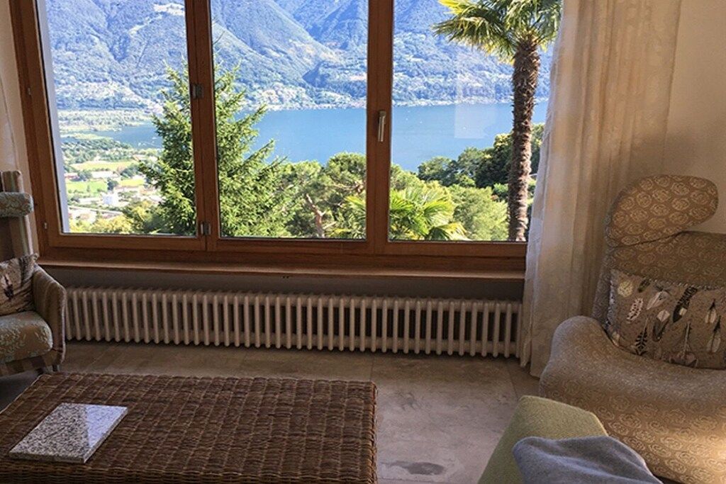 Une pièce dotée d’une grande fenêtre donnant sur un lac et des montagnes, d’un canapé en osier, d’un radiateur et d’un coussin à motifs.