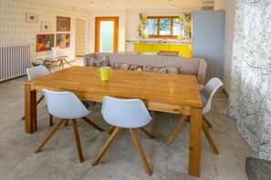 A dining area with a wooden table and white chairs, a sofa, and a kitchen in the background. - Holiday apartment with 1 bedroom (Contra)