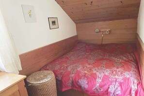 Ein kleines Zimmer mit einem Einzelbett, schrägen Wänden, einem Holz-Kopfstück, einer rot gemusterten Bettdecke, einem kleinen gewebten Hocker und zwei gerahmten Bildern an der Wand.