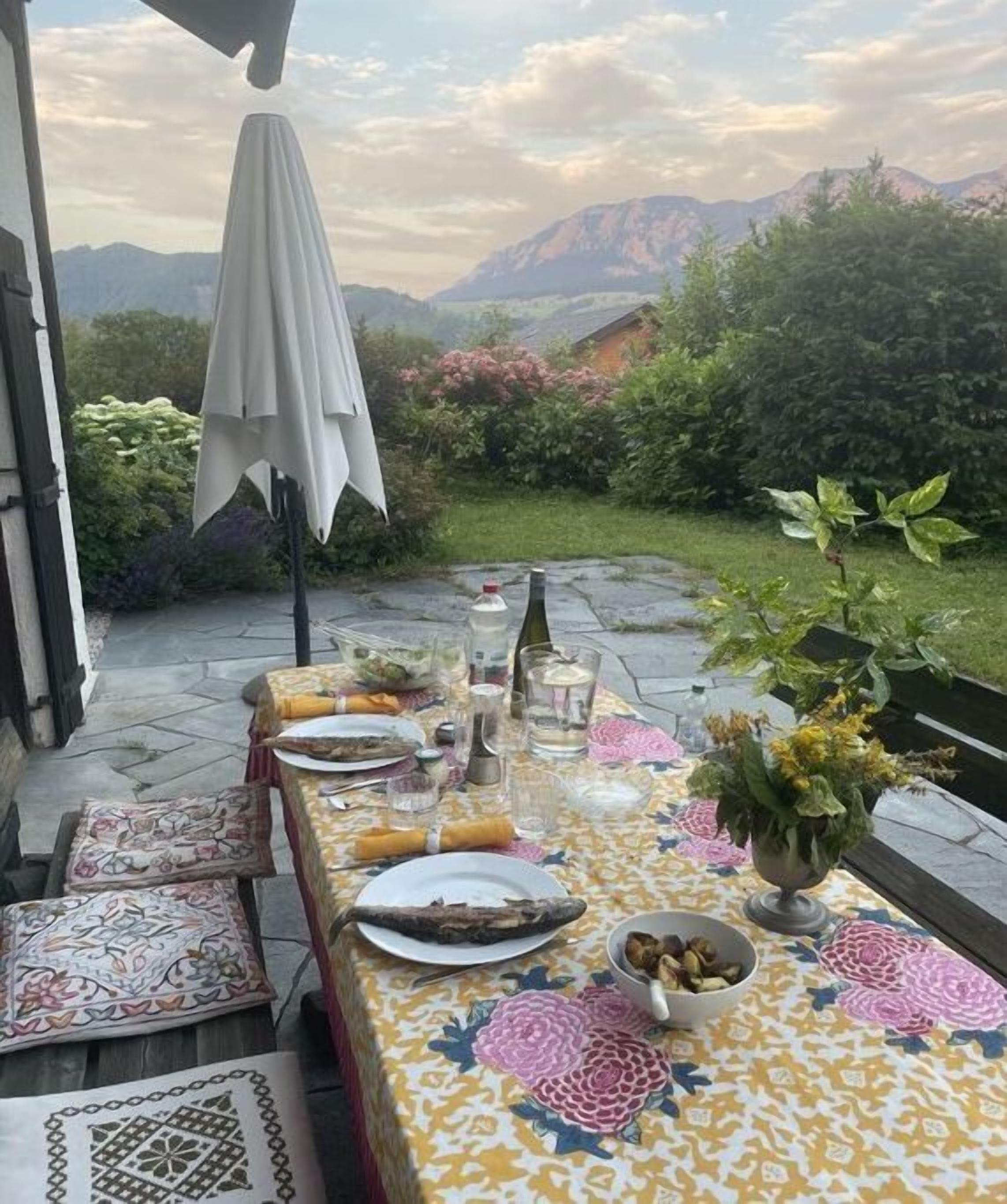 An outdoor dining area with a table set for two, surrounded by a garden with a mountain view.