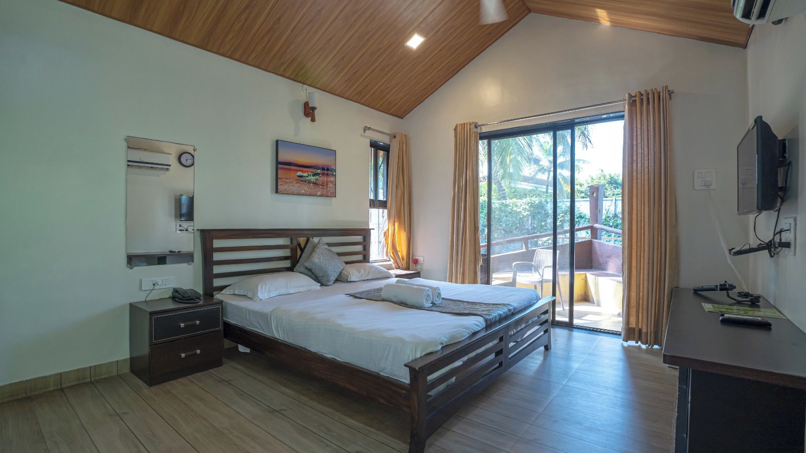 Quarto com cama de madeira, mesa de cabeceira, escrivaninha e uma janela grande com cortinas.