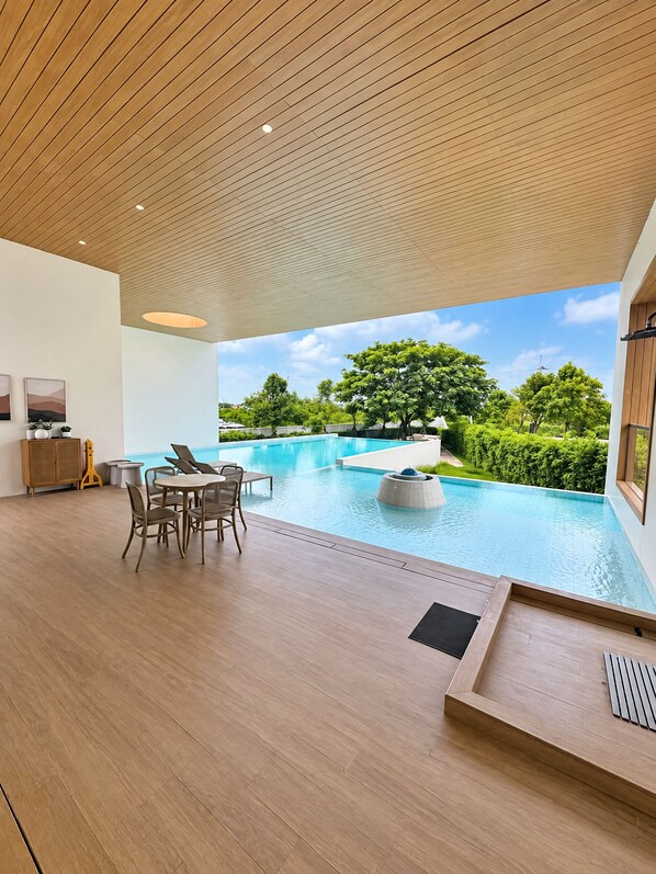 A modern indoor pool area with a wooden ceiling, a seating area with chairs and a table, and a view of a landscaped garden with trees.