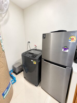 A compact kitchen with a stainless steel refrigerator, a coffee machine, and a wall-mounted fan.