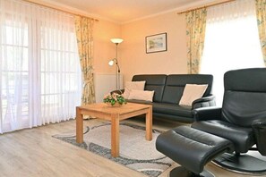 Een woonkamer met een bank, een zwarte leren fauteuil, een houten salontafel en een vloerlamp.