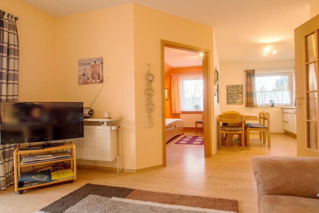 Ein Wohnzimmer mit Fernseher auf einem Schrank, einer Couch, einem Essbereich mit Stühlen und einer Küche im Hintergrund.