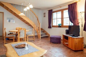 Ein Wohnzimmer mit einer Holztreppe, einem Esstisch mit Stühlen, einem Fernseher auf einem Holzschrank und einem Fenster mit Vorhängen.