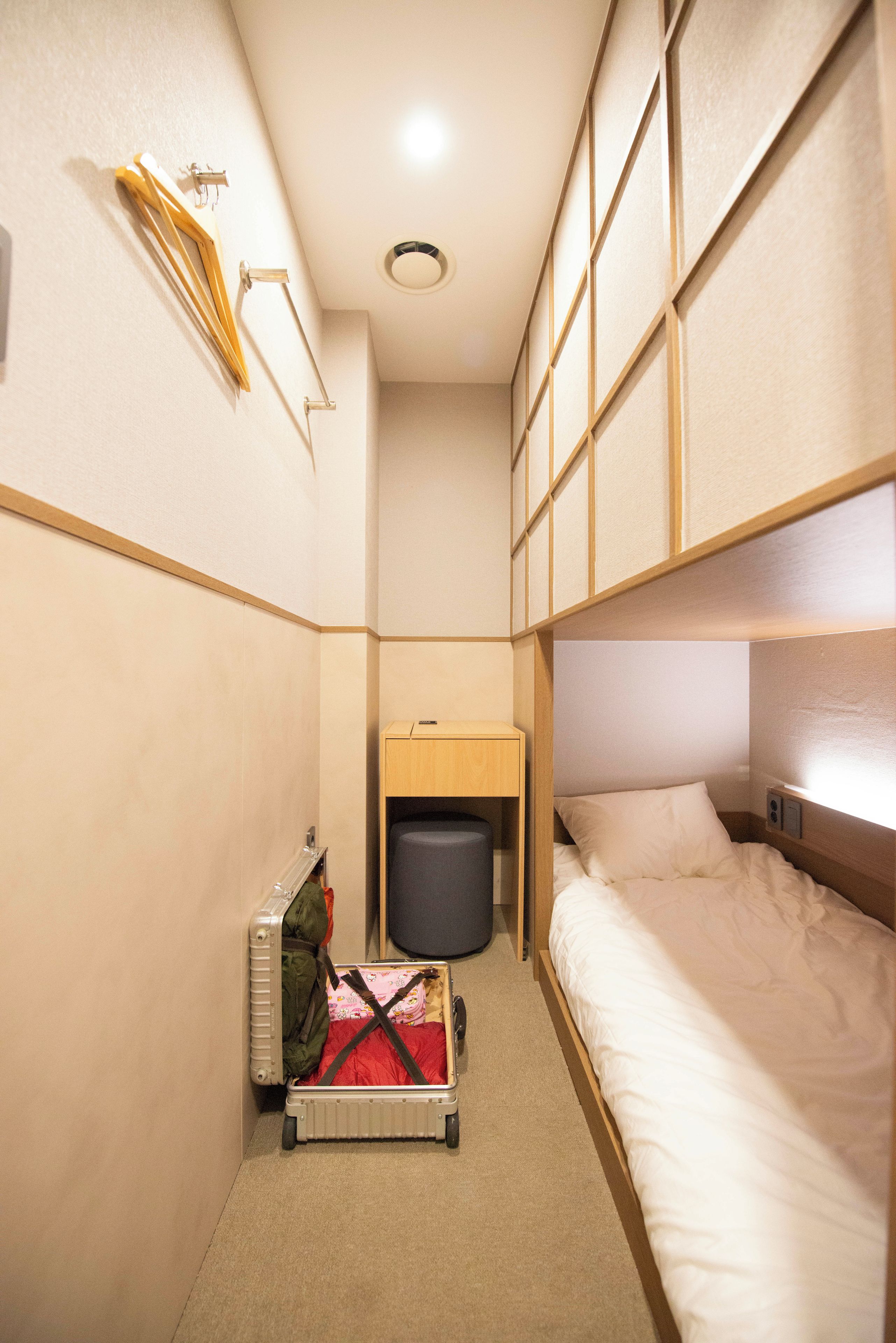 Une chambre d’hôtel compacte avec un lit superposé, un petit bureau, une poubelle et une valise.