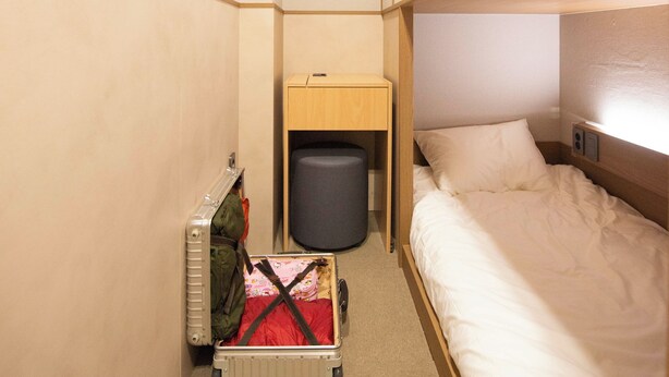 Ett kompakt hotellrum med en våningssäng, ett litet skrivbord, en papperskorg och en resväska.