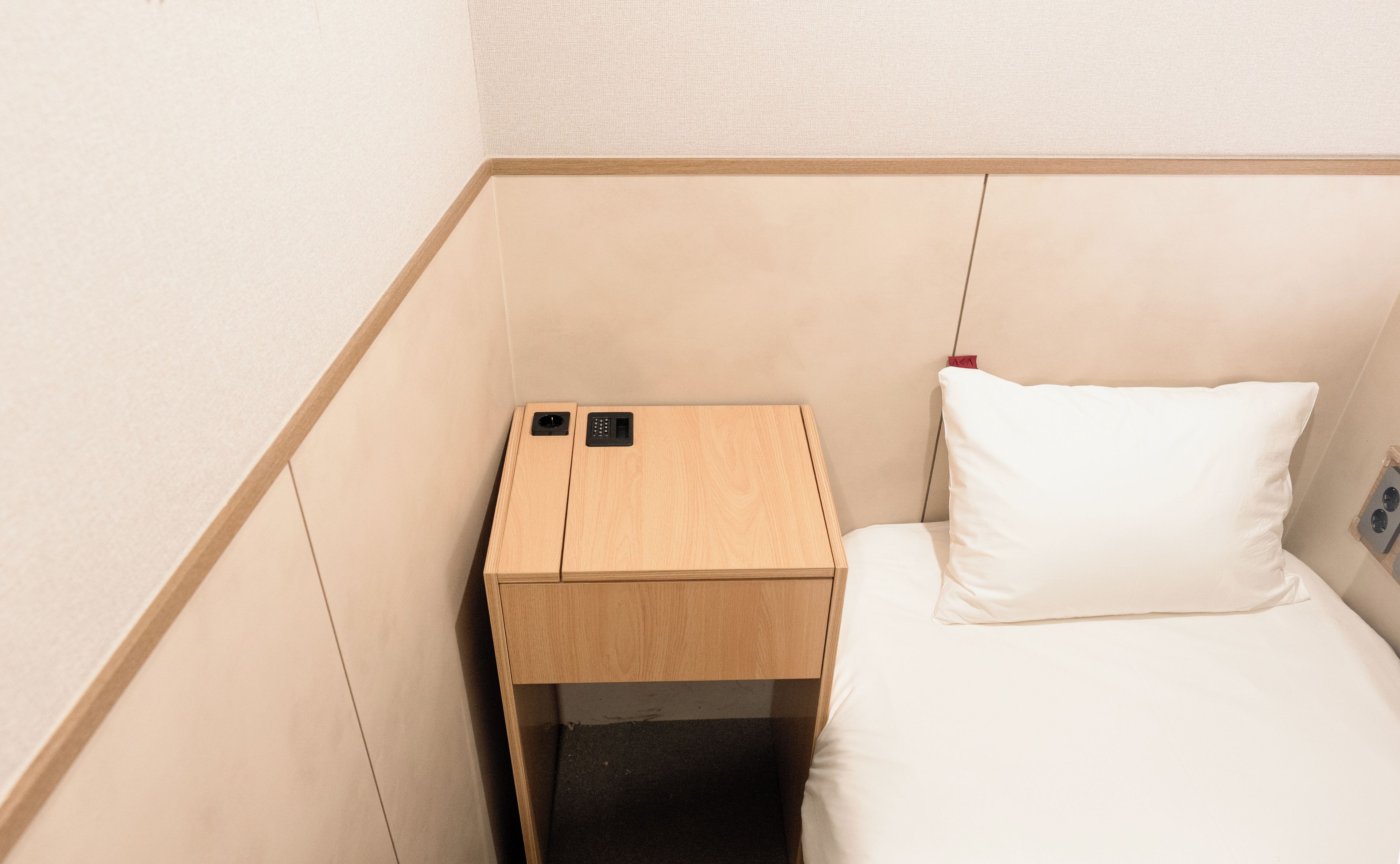 A small, compact room with a wooden nightstand, a bed with a white pillow, and a wall-mounted electrical outlet.