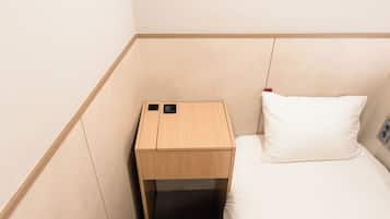 A small, compact room with a wooden nightstand, a bed with a white pillow, and a wall-mounted electrical outlet.
