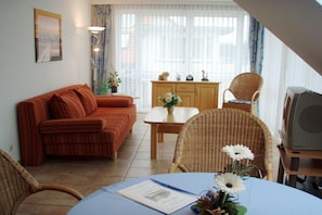 A living room with a sofa, chairs, a table with a vase of flowers, a cabinet, and a television. - Residence on the Beach 1-18 (Zingst (Ostseeheilbad))