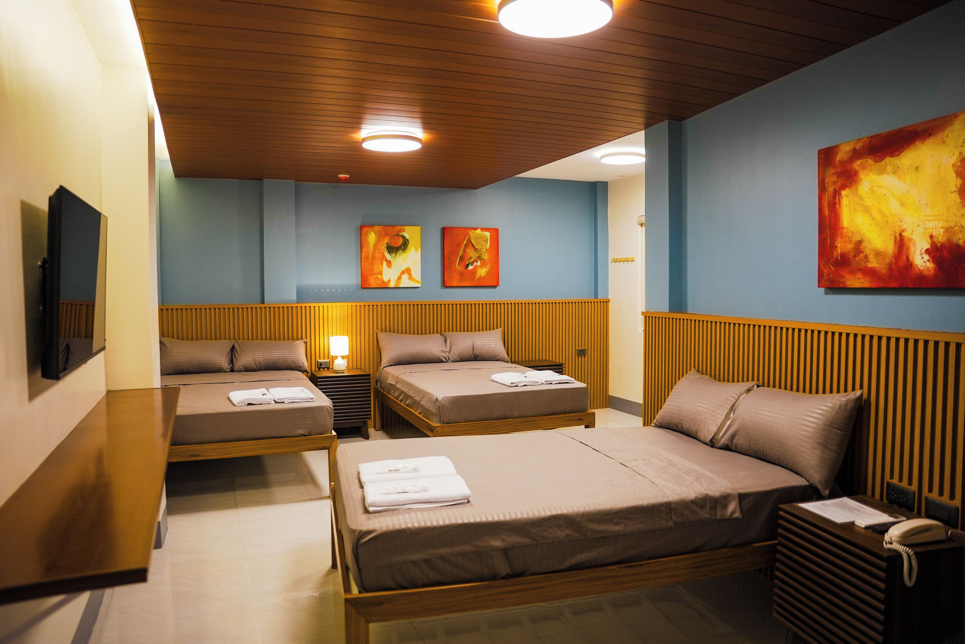 A hotel room with two single beds, a larger bed, a desk with a telephone, and artwork on the walls.