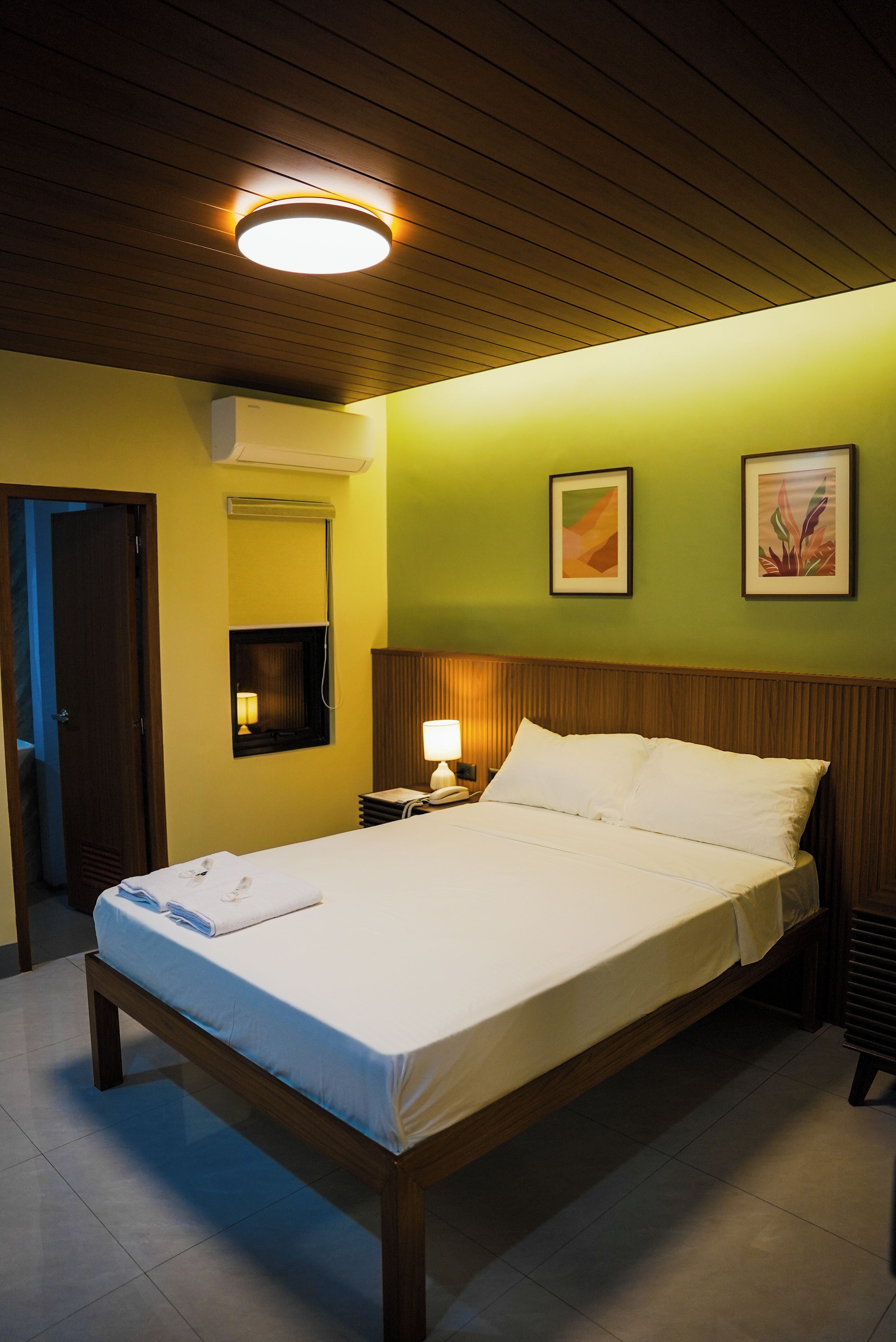 A hotel room with a bed, bedside lamp, wall-mounted air conditioner, and two framed pictures.