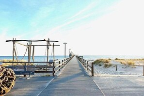 Ein Holzsteg führt zum Strand, links befinden sich eine Bank und ein Mülleimer.