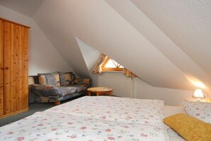 Ein Schlafzimmer mit schrägverdeckter Decke, einem Bett mit floralem Bettzeug, einem Holzschrank, einer Couch mit gemusterten Kissen und einem kleinen Tisch.