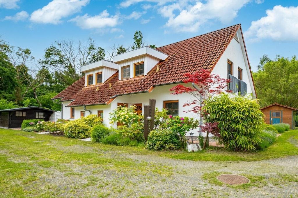 Une maison à deux étages, dotée d’un toit en pente, entourée d’un jardin bien entretenu où poussent différentes espèces d’arbustes, et à laquelle est adossé un petit abri de jardin.