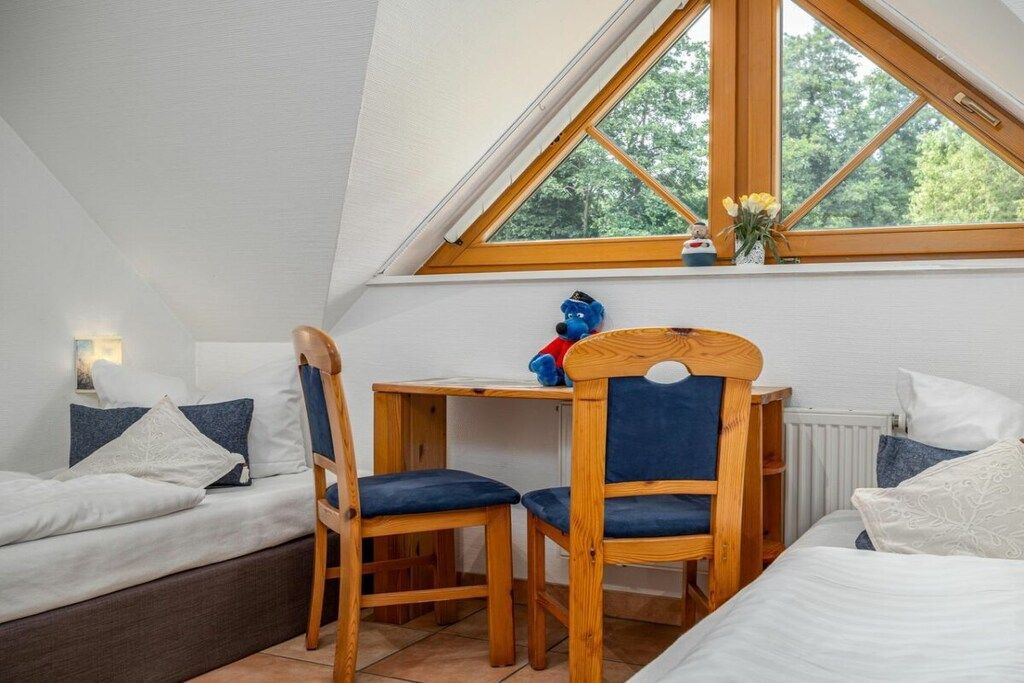 Une pièce avec un plafond en pente, une table en bois avec des chaises, un lit avec une literie blanche et une fenêtre donnant sur des arbres.