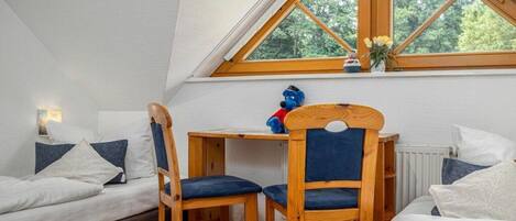 Une pièce avec un plafond en pente, une table en bois avec des chaises, un lit avec une literie blanche et une fenêtre donnant sur des arbres.