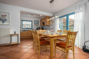 A kitchen with a dining area, featuring a table with chairs, a window with curtains, and a radiator. - 11/T6 holiday home park “Freesenbruch” (Zingst (Ostseeheilbad))