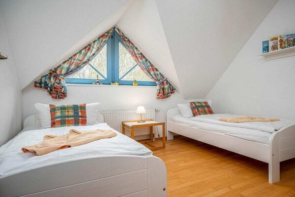 Ett sovrum med två sängar, ett fönster med gardiner, ett litet bord med en lampa och en hylla med böcker.
