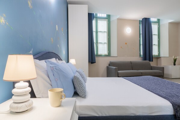 Quarto de hotel com cama, mesa de cabeceira, abajur, xícara de café e sofá.