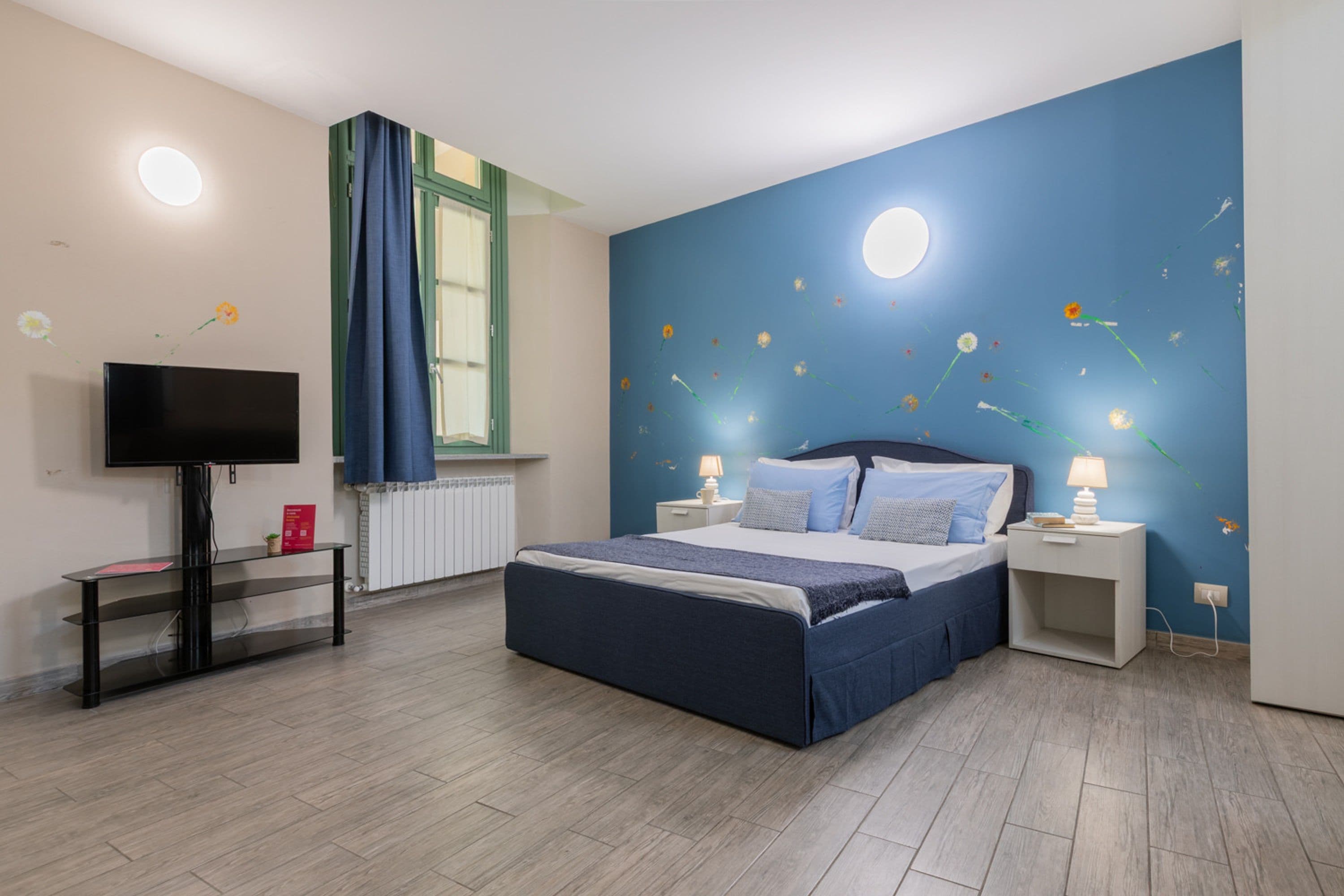 Un dormitorio con cama, mesitas de noche, televisor y ventana con cortinas.