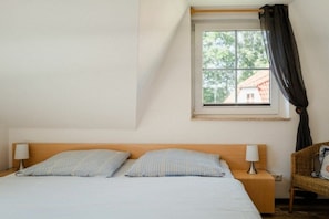 Ein Schlafzimmer mit einem Bett, zwei Nachttischlampen, einem Fenster mit Vorhängen und einem Korbstuhl.