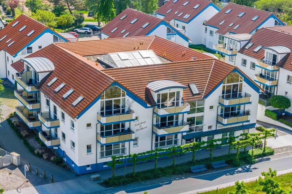 Un immeuble résidentiel à plusieurs étages, doté d’un toit recouvert de tuiles rouges, de balcons et d’un jardin paysager à l’avant.