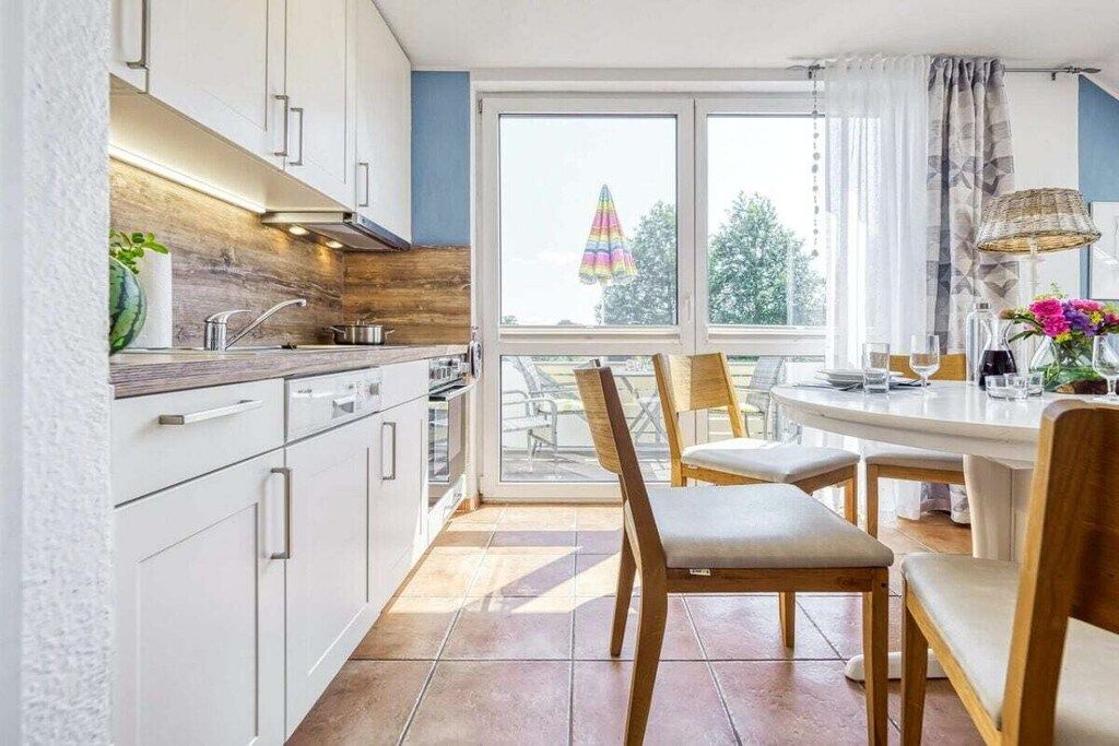 Una cocina moderna con armarios blancos, un ventanal grande y una zona de comedor con sillas de madera.