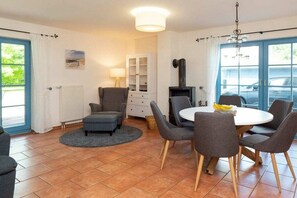A room with a dining table, chairs, a sofa, a cabinet, a fireplace, and a view of the outside. - 11a/T6 holiday home park “Freesenbruch” (Zingst (Ostseeheilbad))