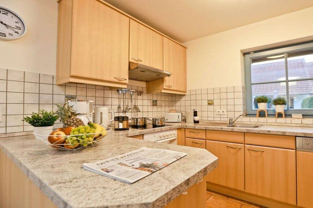 A kitchen with wooden cabinets, a granite countertop, a clock, and a window with plants. - 11a/T6 holiday home park “Freesenbruch” (Zingst (Ostseeheilbad))