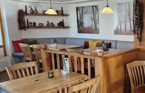 A dining area with wooden tables and chairs, a sofa, and wall-mounted shelves with decorative items. - 11a/T6 holiday home park “Freesenbruch” (Zingst (Ostseeheilbad))