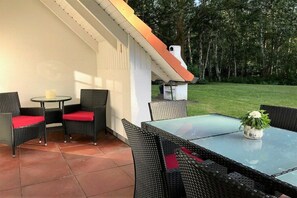 An outdoor patio with wicker furniture and a glass table, surrounded by a white wall and a grassy area. - 11a/T6 holiday home park “Freesenbruch” (Zingst (Ostseeheilbad))