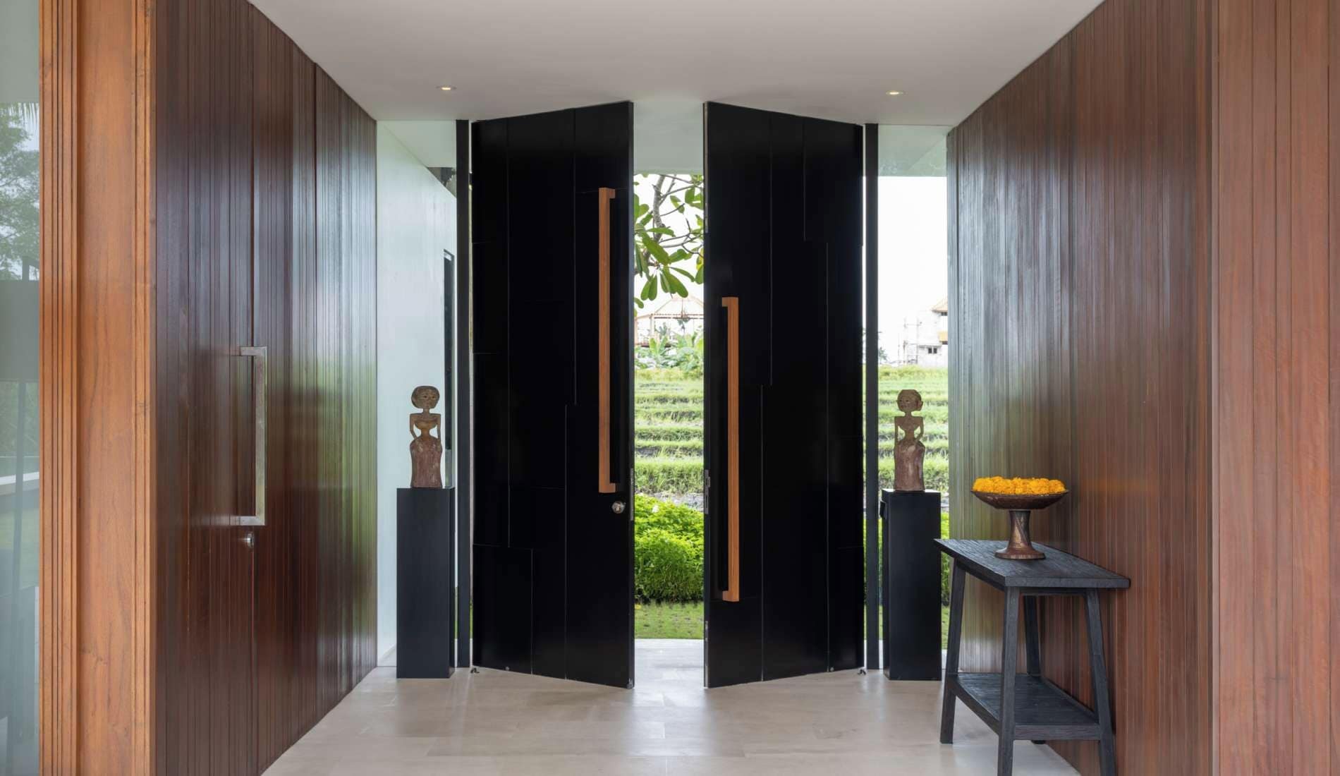 A modern interior with a central open doorway leading to a garden, flanked by wooden panels and two small sculptures on pedestals.