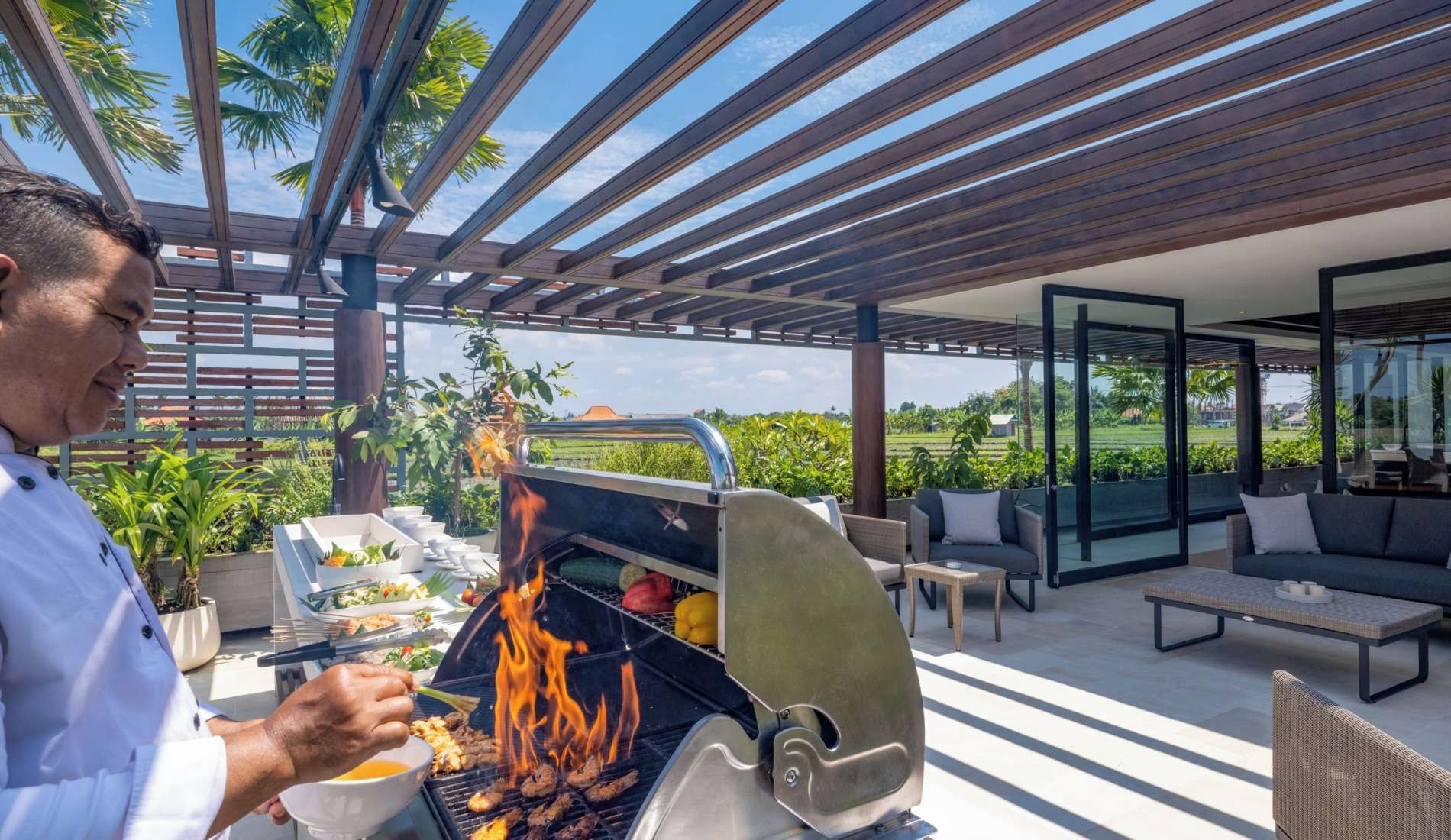 An outdoor grilling area with a modern pergola, a dining setup with a sofa and table, and a view of the surrounding greenery and landscape.