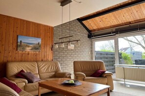 A room with a wooden ceiling, a brick wall, a wooden floor, a beige sofa, a wooden coffee table, and a window with a view of the outside. - Holiday home with 3 bedrooms (LB Zoutelande)