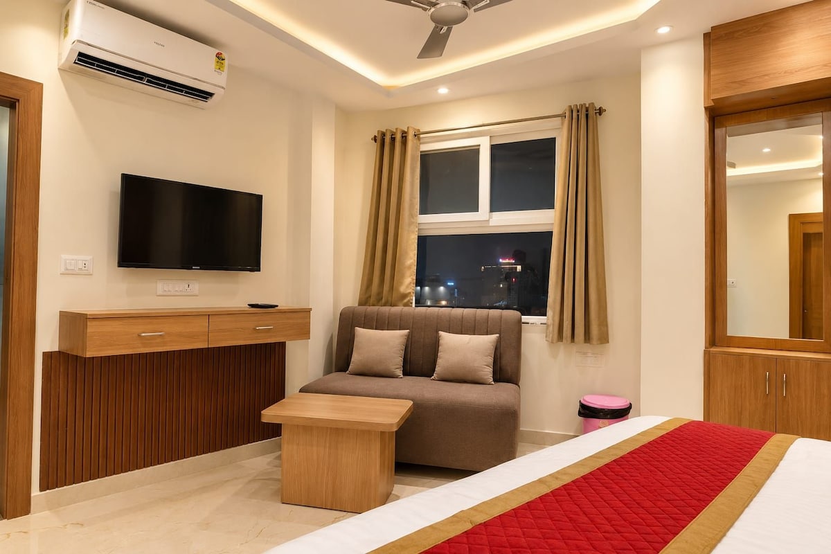 Una habitación de hotel moderna con una televisión de pantalla plana, un sofá, una mesita de madera, una cama con colcha roja y blanca, y una ventana con cortinas.