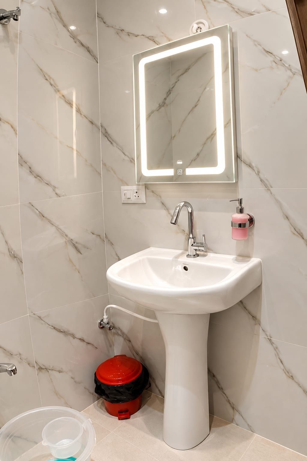Un baño con lavamanos pedestal blanco, un espejo con iluminación integrada, un cubo de basura rojo y un recipiente de plástico transparente.