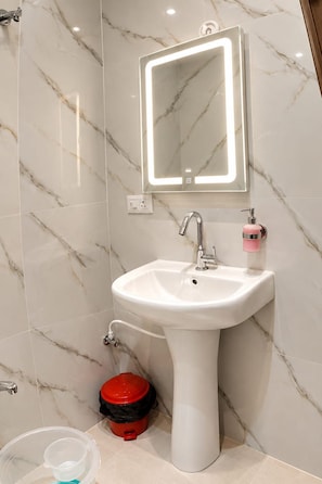 Un baño con lavamanos pedestal blanco, un espejo con iluminación integrada, un cubo de basura rojo y un recipiente de plástico transparente.