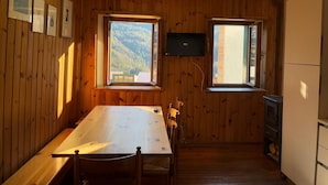 A wooden-paneled room with a dining table, chairs, a television, and two windows offering a view of a mountainous landscape. - Casa Le Triestine: cozy, traditional Dolomite accommodation for groups (Venas di Cadore)