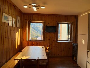Ein Zimmer mit Holzwänden, ausgestattet mit einem Esstisch, Stühlen, einem Fernseher und zwei Fenstern, die einen Blick auf eine gebirgige Landschaft bieten.