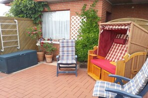 Eine Terrasse mit einem Korbstuhl und gepolstertem Sitz, einer Holzbank und einer Leiter an einer Ziegelwand.