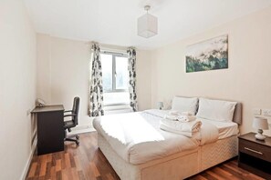 A bedroom with a bed, a desk, a chair, a window with curtains, a painting on the wall, and a bedside table with a lamp. - Gorgeous 2BR Flat 0.1 mi ExCel & Prince Regent St. (London)
