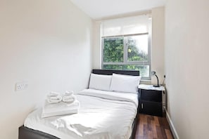 A bedroom with a bed, a nightstand, a window, and a wall socket. - Gorgeous 2BR Flat 0.1 mi ExCel & Prince Regent St. (London)