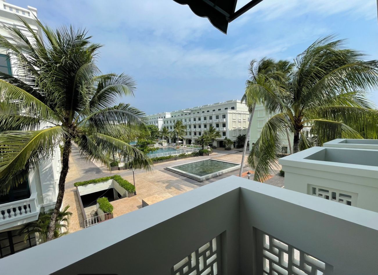 Vista de uma varanda com vista para uma área de piscina, palmeiras e um edifício ao fundo.