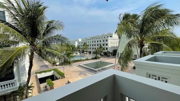 Vista de uma varanda com vista para uma área de piscina, palmeiras e um edifício ao fundo.
