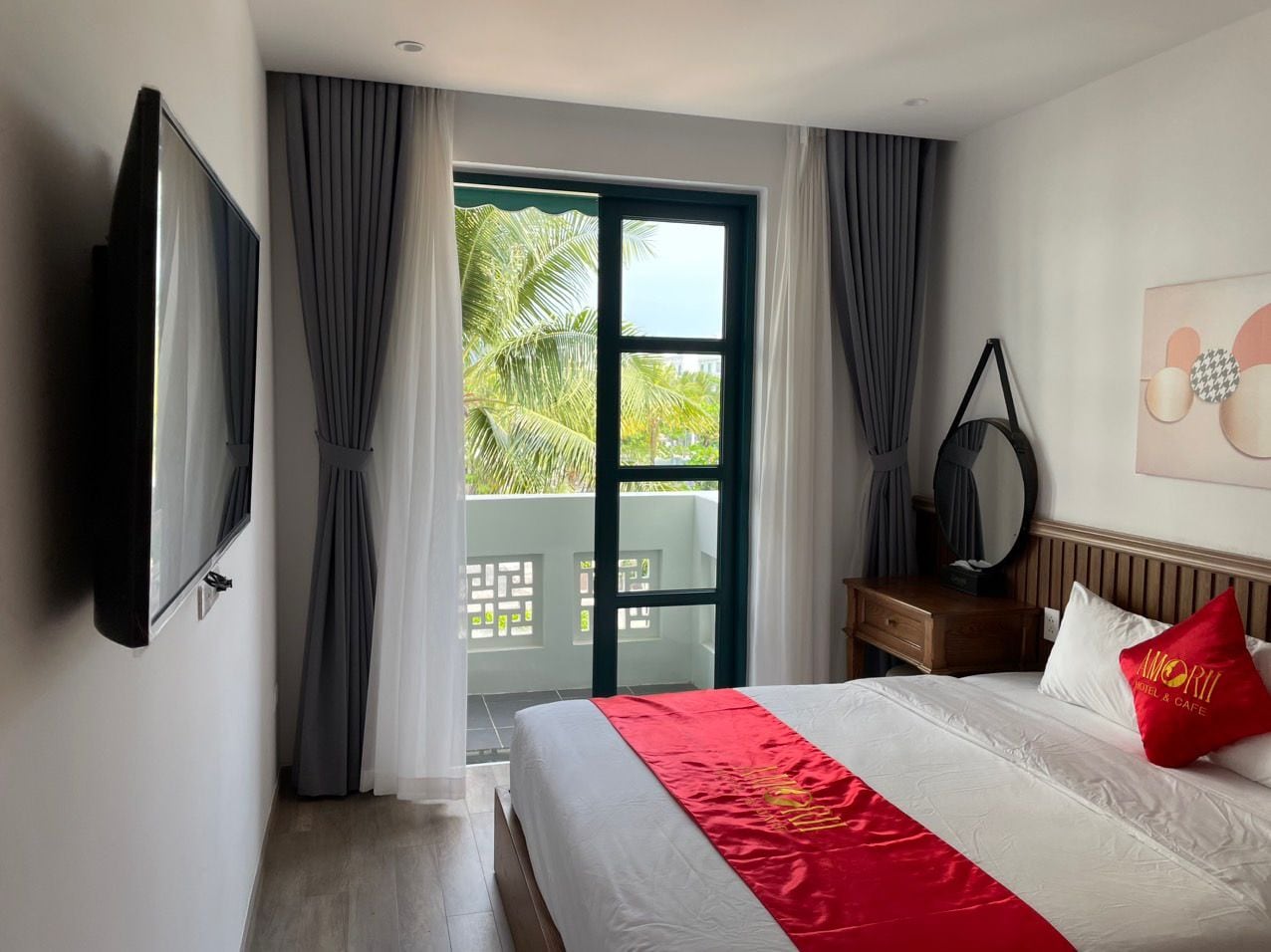 A hotel room with a bed, a television, a balcony with curtains, and a view of greenery.