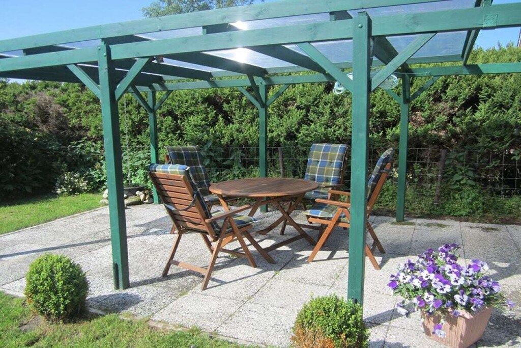 Un patio al aire libre con una mesa y sillas de madera, rodeado por una estructura de pérgola verde.